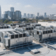 The image shows multiple commercial rooftop HVAC units positioned atop a large office building, with a bustling city skyline in the background